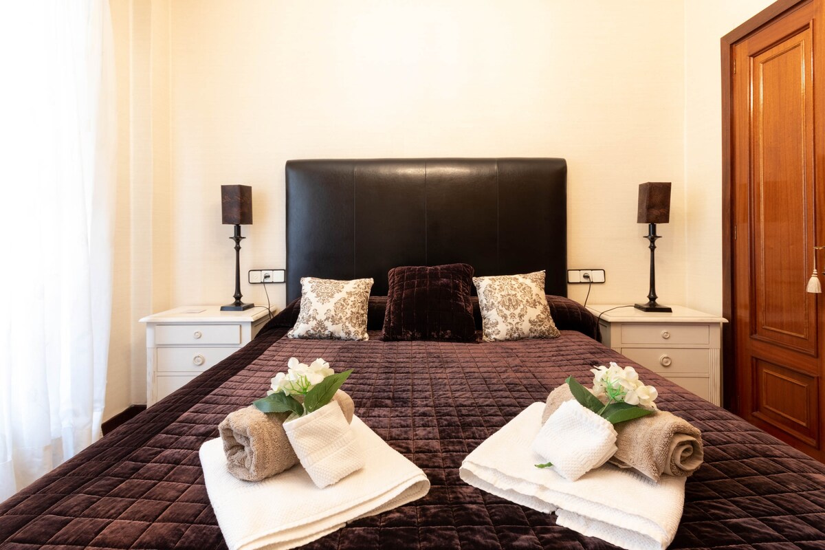 A spacious sleeping area features a large dark headboard complemented by bedside tables. Two decorative pillows rest against the headboard, while neatly rolled towels adorned with flowers are placed at the foot of the bed. Natural light filters through a nearby window.