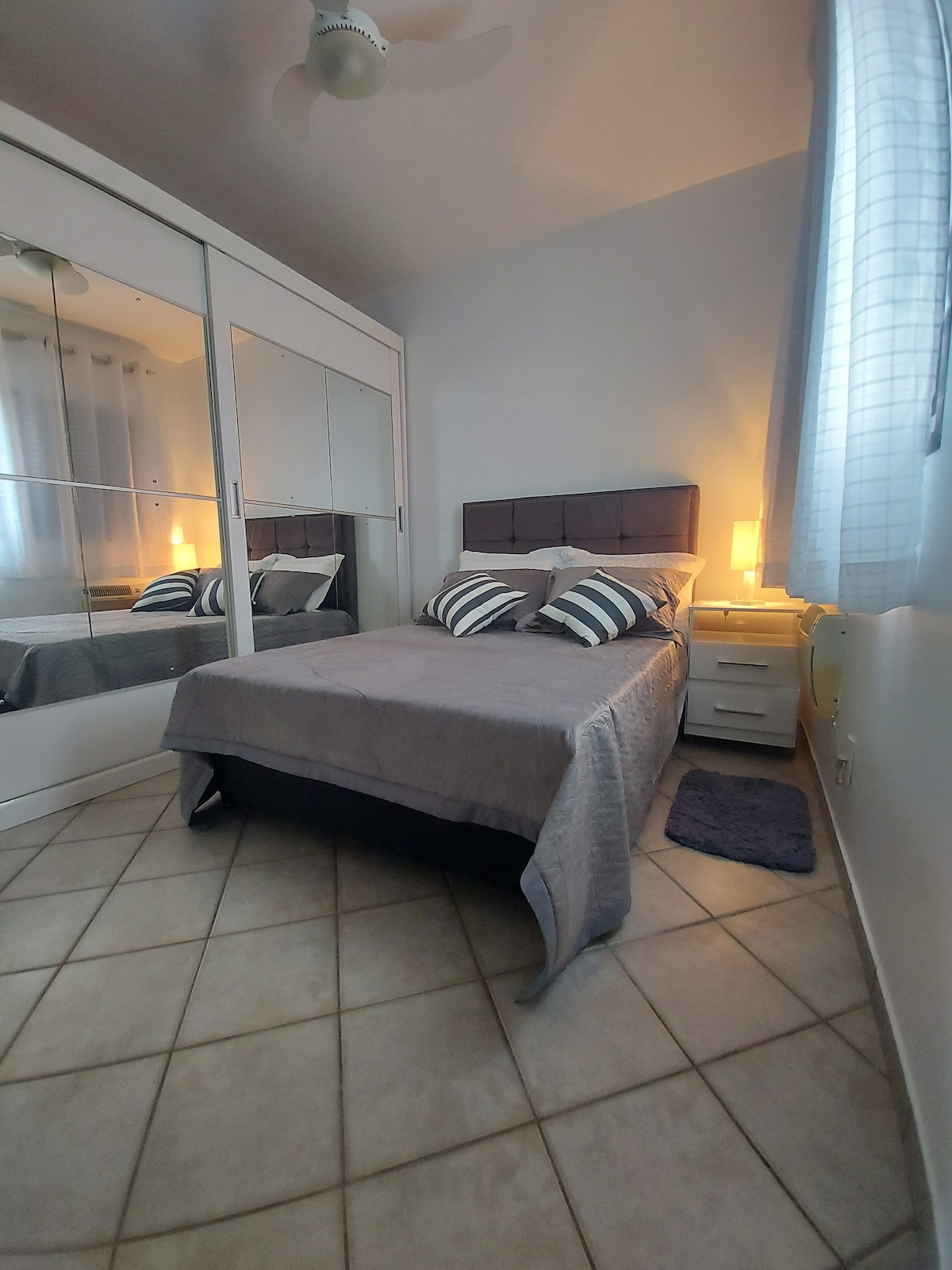 A well-lit bedroom features a spacious bed with decorative pillows, flanked by bedside tables. A mirrored wardrobe reflects the room's soft lighting, and a small rug lies beneath the bed. Lace curtains allow natural light to filter through a nearby window.