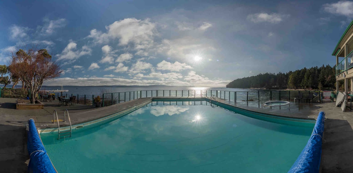 An expansive outdoor pool is visible, reflecting the sky and surrounding landscape. Clear waters are highlighted by the sun's reflection, while a scenic backdrop features trees and the ocean. The area includes a hot tub and lounge space for relaxation.