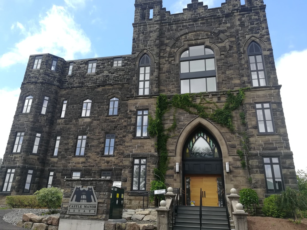 A historic stone building features a grand arched entrance framed by intricate architectural details. Large windows are adorned with greenery, and the structure's rugged exterior is complemented by surrounding landscaping. A sign indicating 'Castle Manor' stands prominently at the entrance.