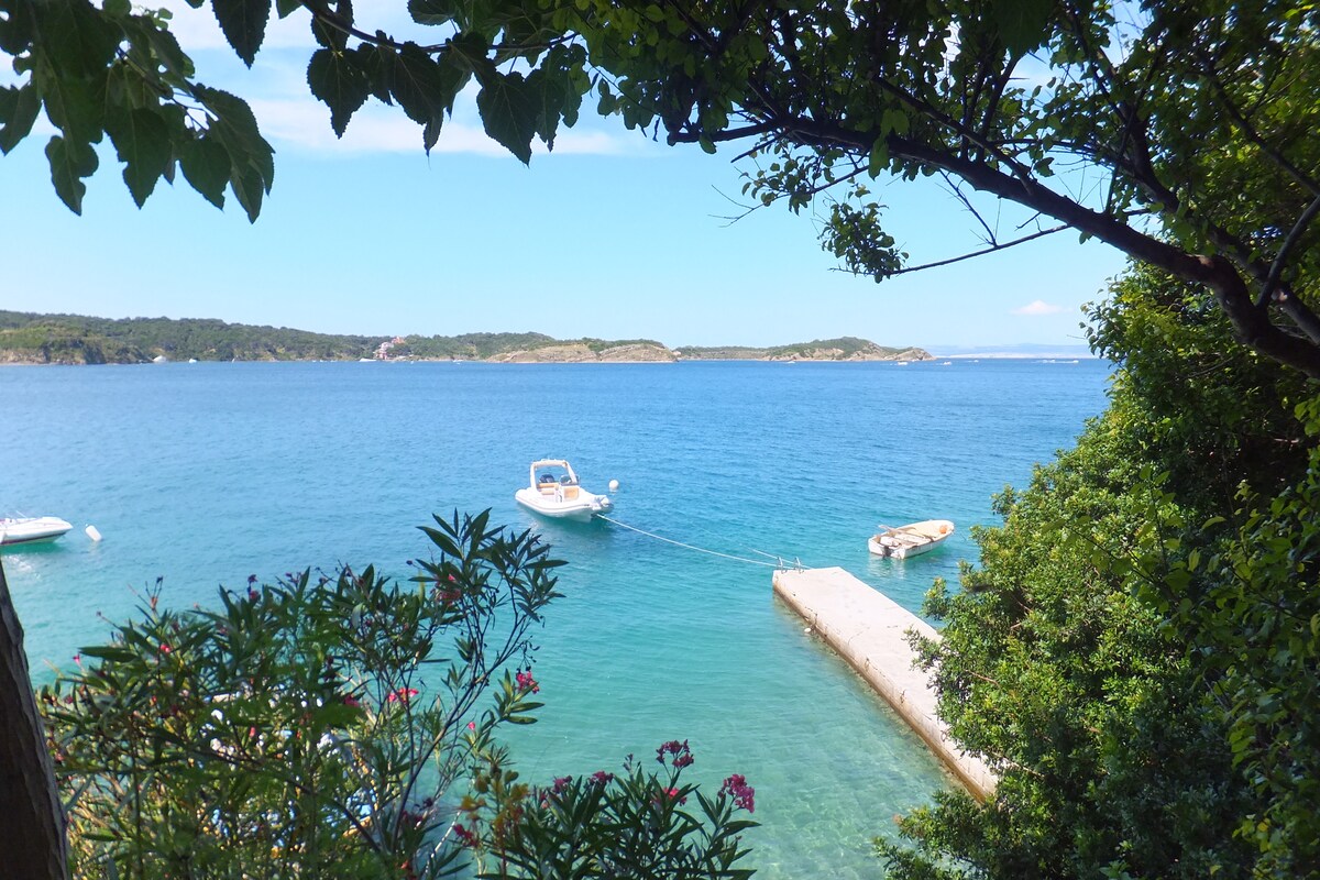 A serene waterfront view is framed by lush greenery, showcasing clear blue waters and moored boats. The shoreline features a private dock, inviting leisure and relaxation. The landscape includes distant islands and hills, enhancing the tranquil atmosphere of the coastal environment.