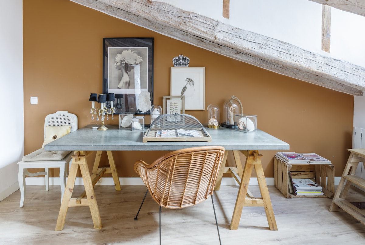 A spacious work area features a large table supported by wooden trestles, complemented by a rattan chair. The room is accented by a warm brown wall, and decorative items are arranged on the table, including framed artwork and lamps.