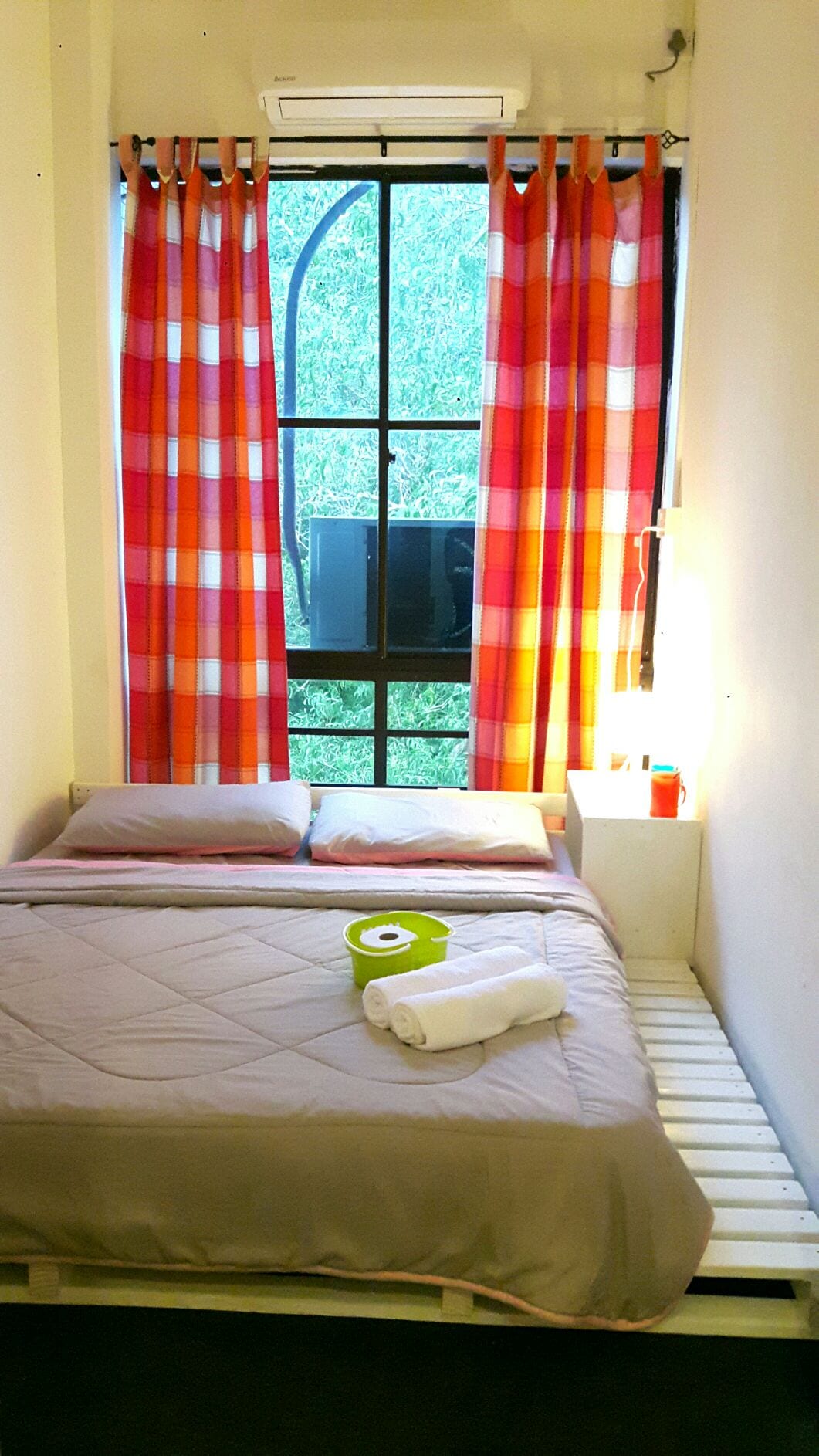 A cozy double room features a pallet bed covered with a simple quilt. Brightly colored curtains frame a large window, allowing natural light to illuminate the space. A small bedside table holds a bowl and towels, while a modern air conditioning unit is mounted above.