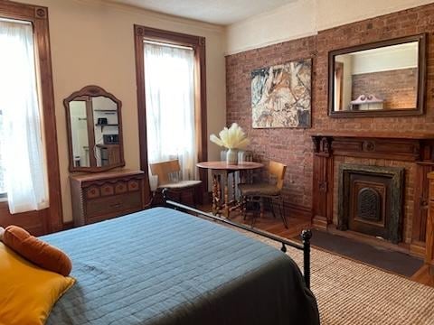 A spacious studio features exposed brick walls and hardwood flooring. Natural light filters through the two tall windows with sheer curtains. A cozy queen-sized bed is positioned across from a petite dining table with two chairs. A vintage dresser and a decorative mirror add character to the space.