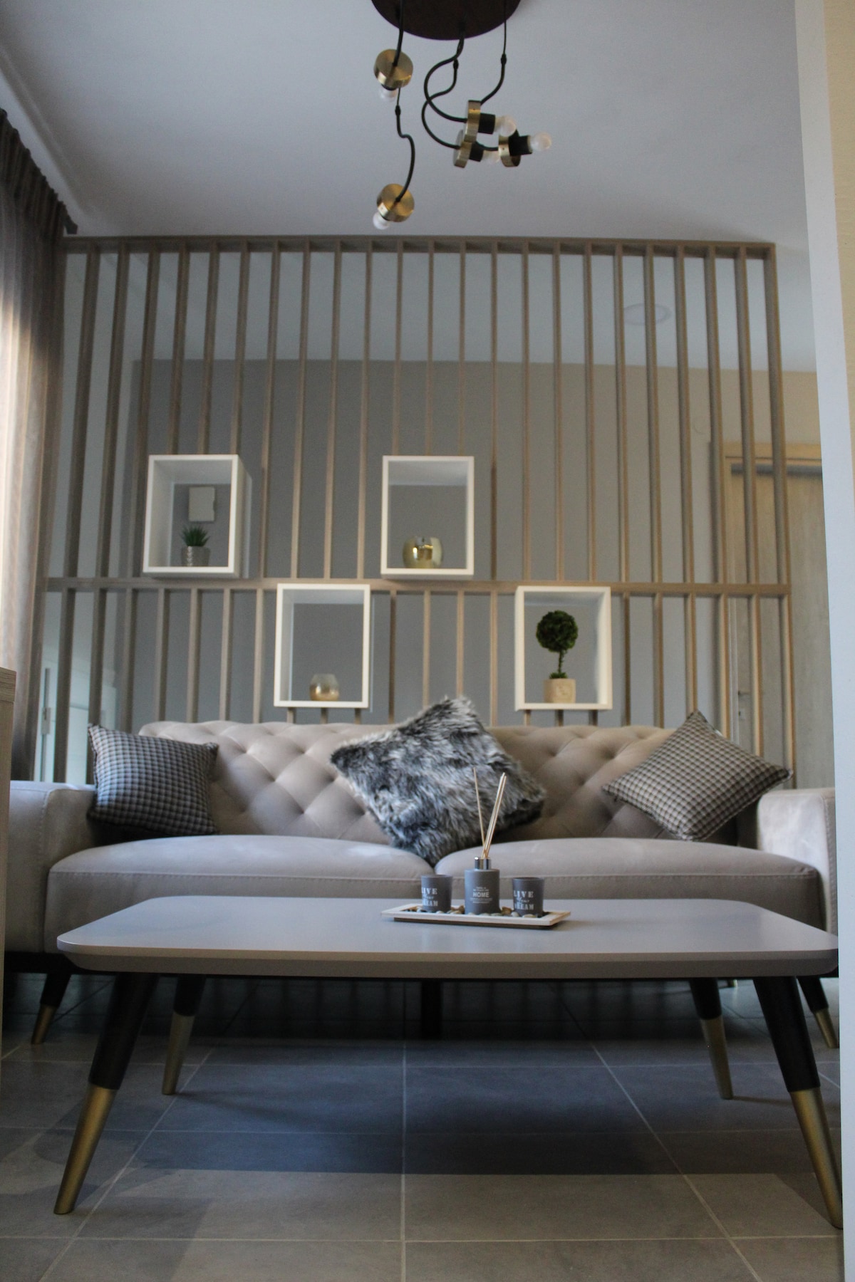 A modern living area features a tufted sofa adorned with decorative pillows. A sleek coffee table is positioned in front of the sofa, displaying a small decorative tray. The backdrop includes a decorative lattice and wall-mounted shelves with potted plants.