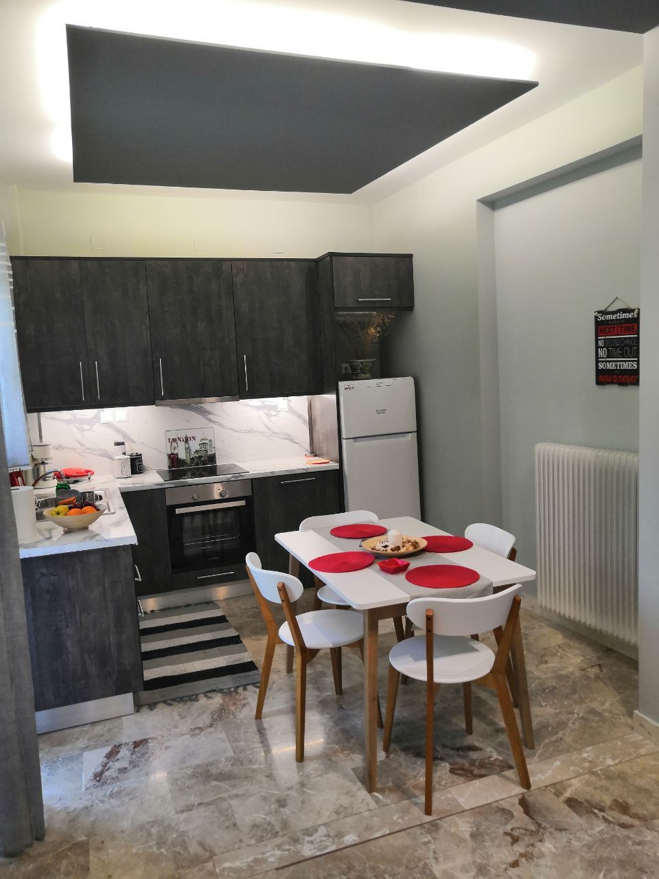 A modern kitchen area includes dark cabinetry and a white marble backsplash. A small dining table with white chairs is set for four, featuring red placemats. Appliances such as a refrigerator and oven are visible, alongside various kitchen items arranged on the counter.