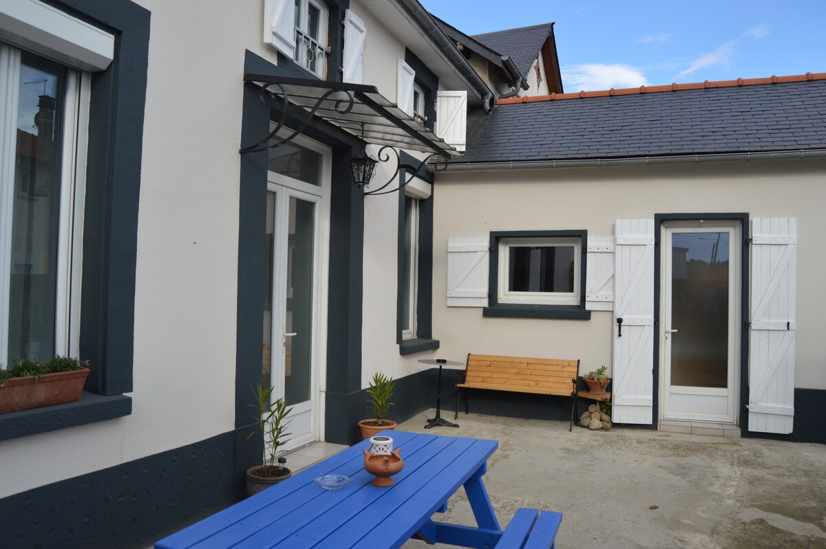 An outdoor space features a blue table, surrounded by potted plants. A wooden bench is positioned against the wall, while white shutters frame the windows. The entrance to the house is visible with a door leading into the interior, and an awning provides shade above.