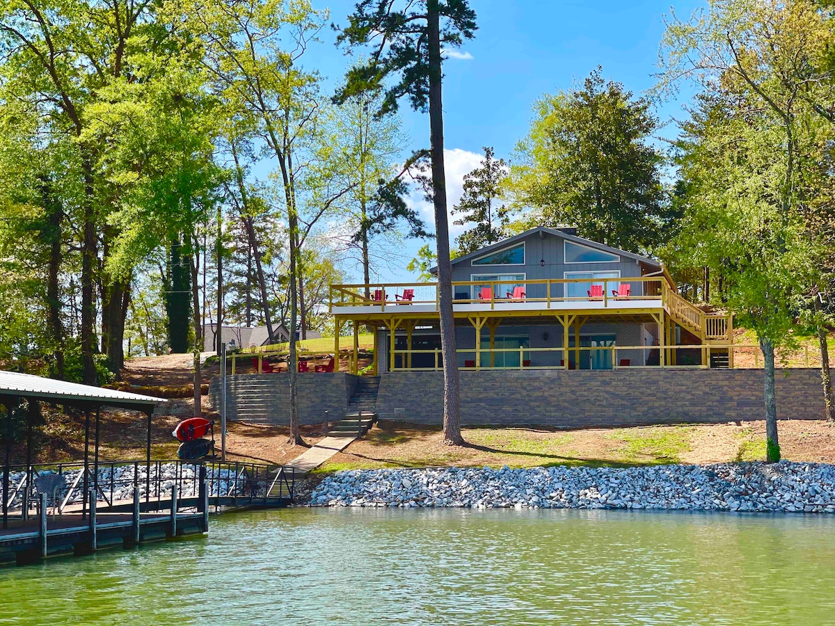 The exterior of a modern cabin is displayed, featuring a spacious upper deck with vibrant red chairs. The cabin sits on a slope amidst lush green trees, overlooking the serene water of Lake Hartwell. A covered dock is visible nearby, enhancing the waterfront appeal.