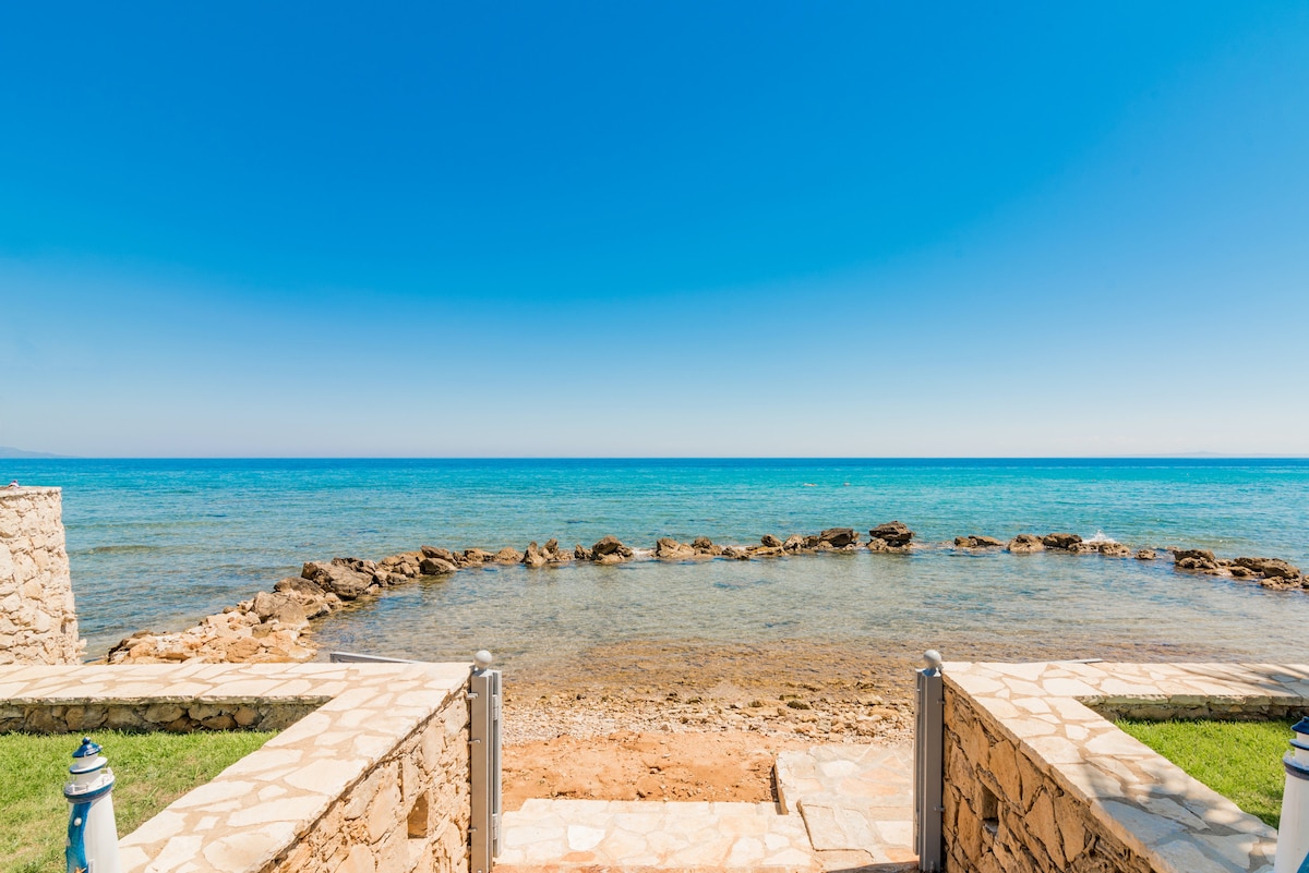 The image captures a serene coastal view with clear blue water extending to the horizon. Rocky formations emerge from the water, creating a natural border. A stone pathway leads to the shore, accompanied by well-maintained grassy areas on either side, enhancing the tranquil seaside atmosphere.