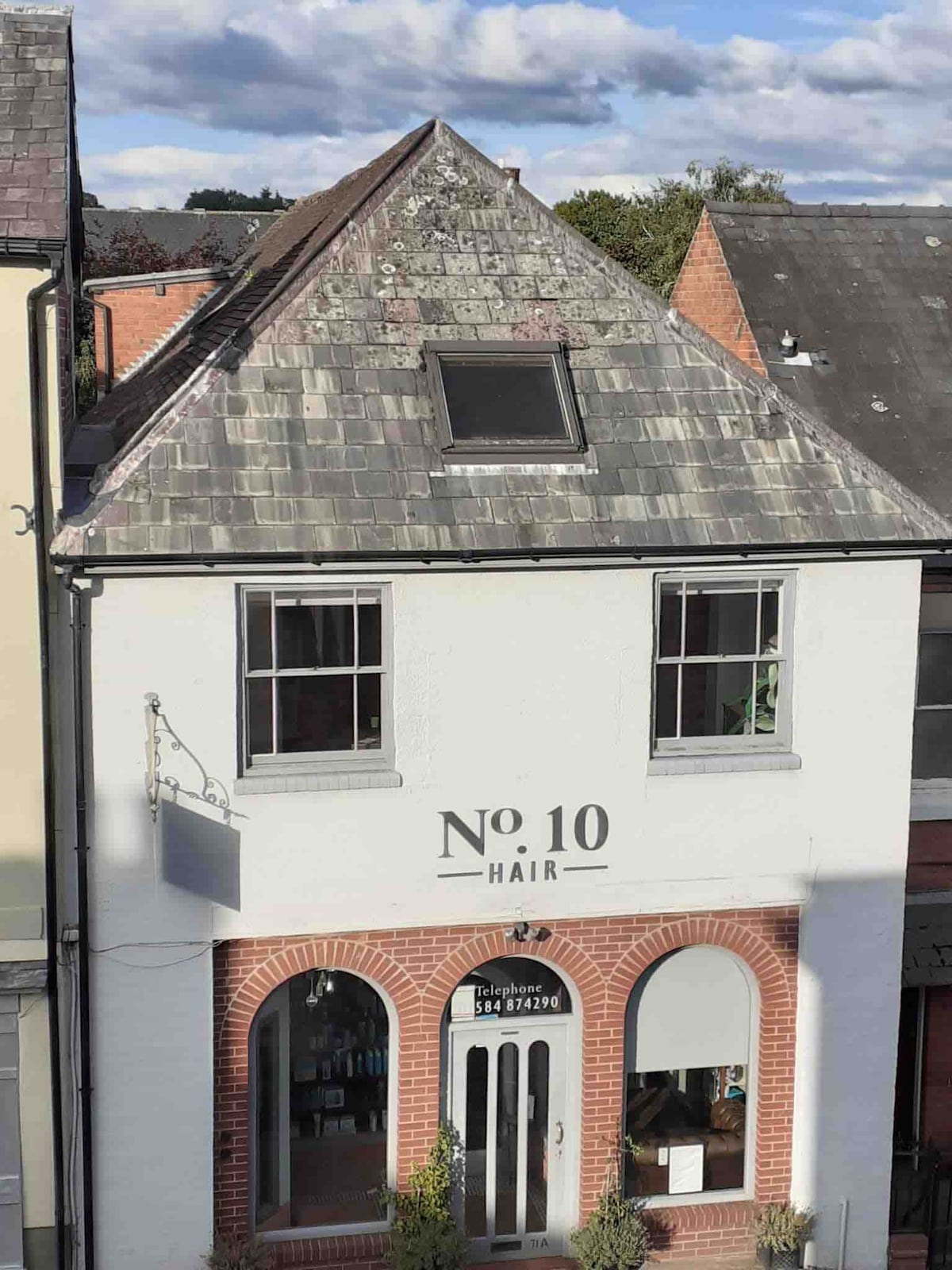 The exterior of a historic building features a white façade with a large window displaying a hair salon. The sloped roof is covered with dark shingles, and a small skylight is visible. The signage above the entrance includes the phrase 'No 10 HAIR'.