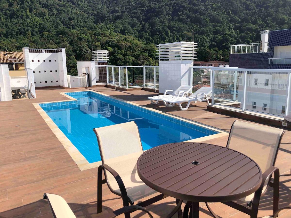 A rooftop area features a modern swimming pool surrounded by wooden decking. Two lounge chairs are positioned near the pool, and a round table with a single umbrella sits nearby, offering a relaxing space with views of lush greenery.