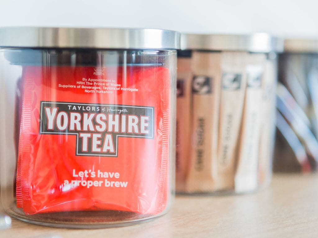 A transparent jar filled with red packaging of Yorkshire Tea is prominently displayed in the foreground. Behind it, additional jars are visible, containing assorted tea bags and sugar packets. The items are arranged on a light wooden surface.
