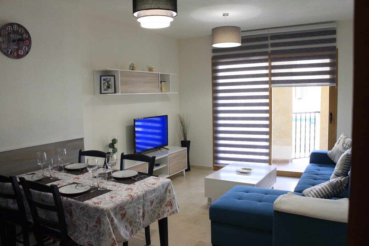 A modern living area is depicted with a blue sectional sofa and a white coffee table. The dining table is set for four with glassware arranged. A television is positioned on a light wood stand, complemented by decorative elements on the shelves. Natural light filters through striped blinds.