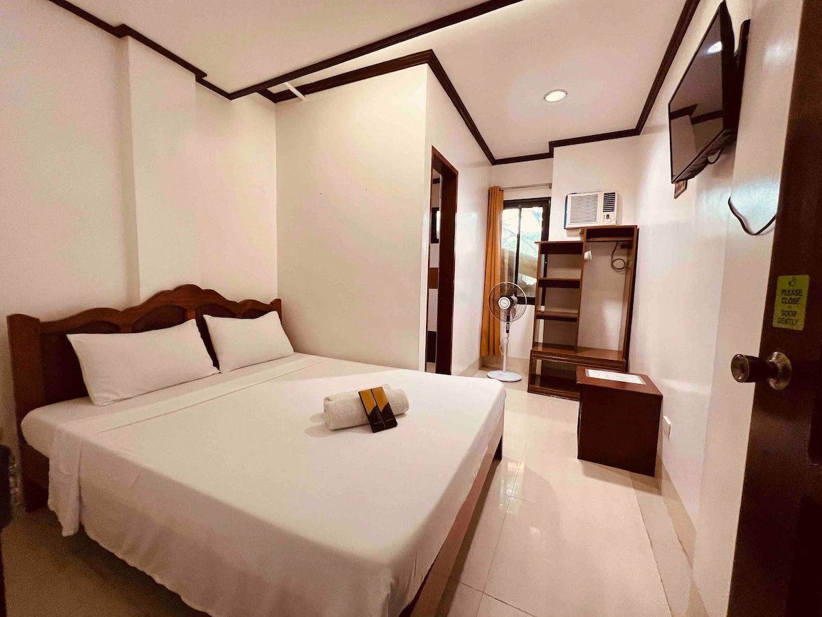 A bright room features a spacious bed with white linens and a neatly arranged towel on top. Dark wood furniture includes a hanging cabinet and a bedside table. A flat-screen television is mounted on the wall, and light enters through a window draped with sheer curtains.