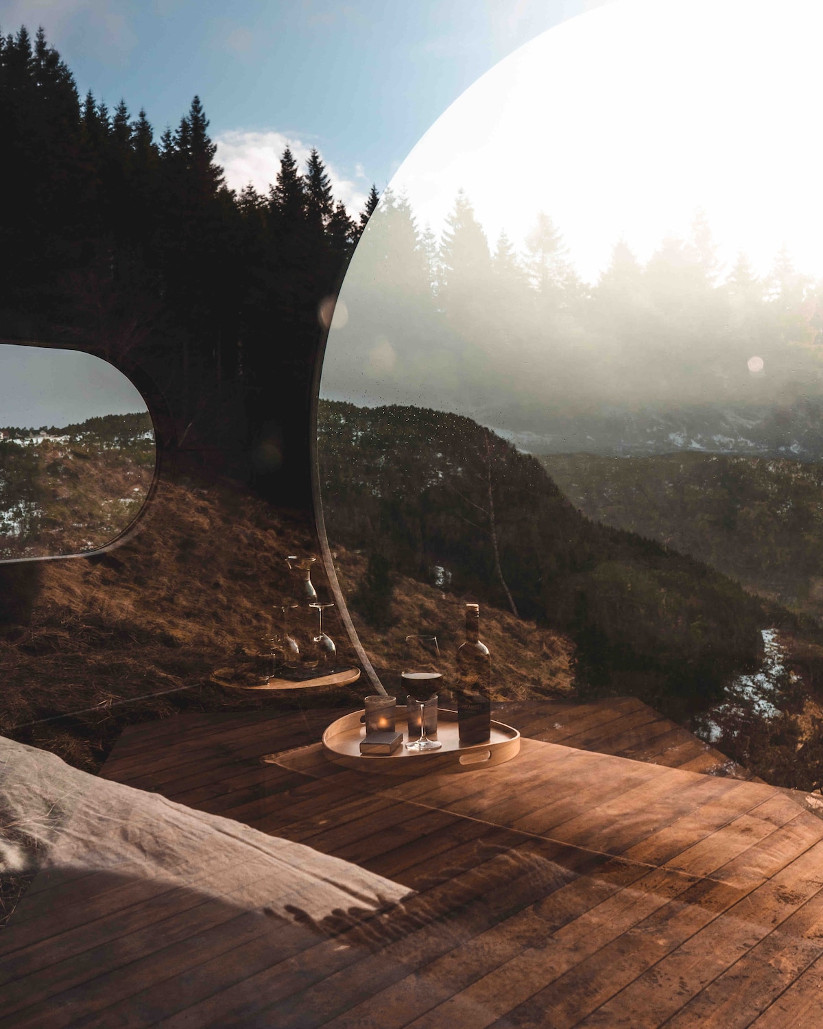 A wooden deck is showcased with a round tray holding glasses and a bottle, overlooking a serene landscape of trees and hills. Soft sunlight filters through, creating a warm ambiance while reflecting off the glass surface, inviting moments of relaxation.