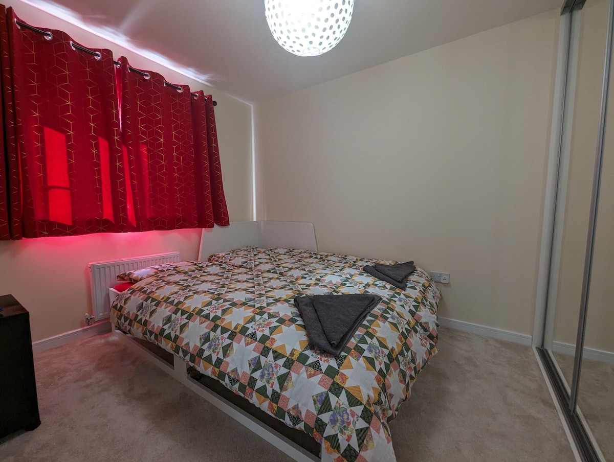 A bright bedroom features a spacious bed covered with a colorful quilt. Red curtains filter natural light, and a geometric patterned ceiling light provides additional illumination. A built-in wardrobe is visible, reflecting a modern design. A neutral carpet adds to the room's comfort.