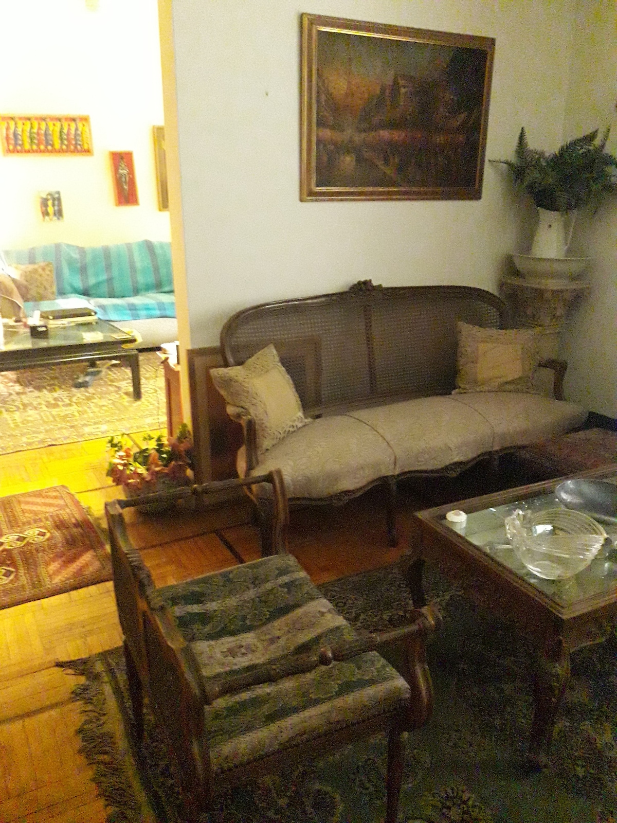 A vintage sitting area features a delicate sofa with woven armrests, complemented by two wooden chairs with patterned upholstery. A glass coffee table rests on a decorative rug, while a wall-mounted painting adds visual interest. Natural light brightens the scene from an adjacent room.