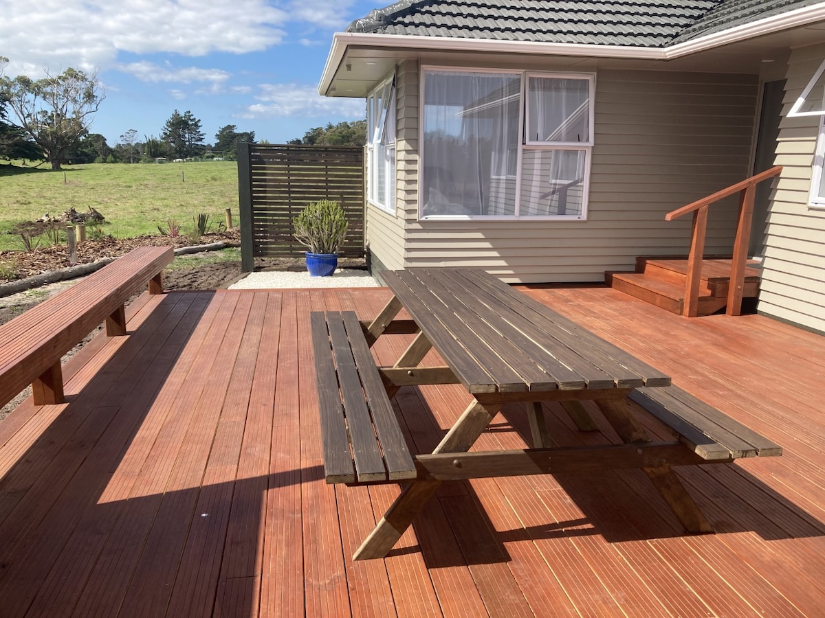 A spacious outdoor deck is featured, equipped with a large wooden picnic table and benches. Sunlight illuminates the area, highlighting the deck's rich, warm tones. A planter with greenery can be seen near the side of the house, with an expansive grassy landscape in the background.