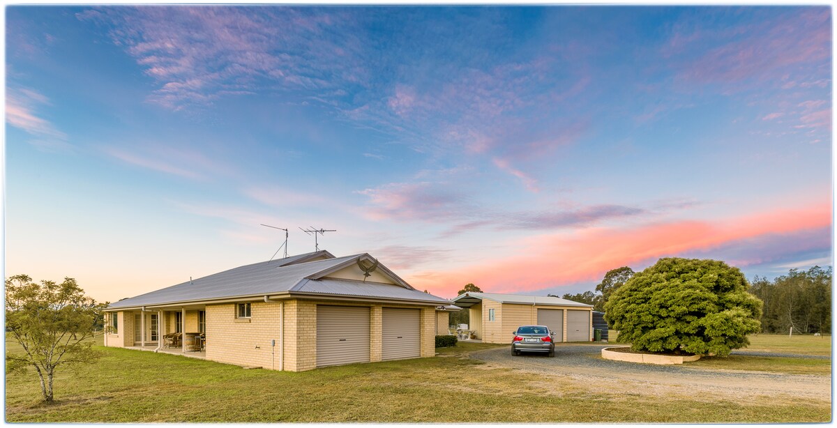 A spacious freestanding cottage is set against a colorful sunset sky. The exterior features warm beige brick walls and a grey roof. Nearby, a gravel driveway leads to a parked car, with open grounds and greenery on display in the foreground.