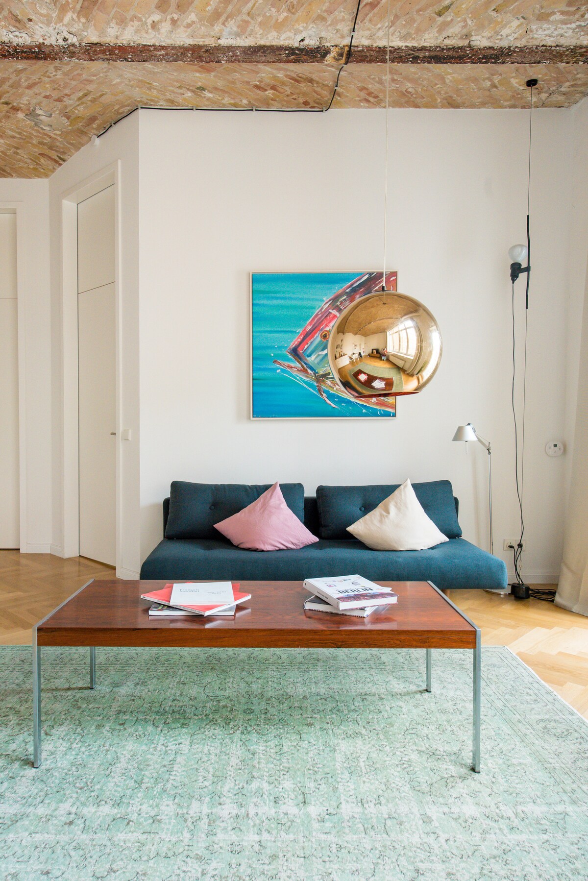 A bright living area features a comfortable sofa adorned with decorative cushions. A sleek coffee table sits on a textured rug, with magazines casually displayed. A colorful artwork hangs on the wall, while a unique pendant light adds visual interest to the space.
