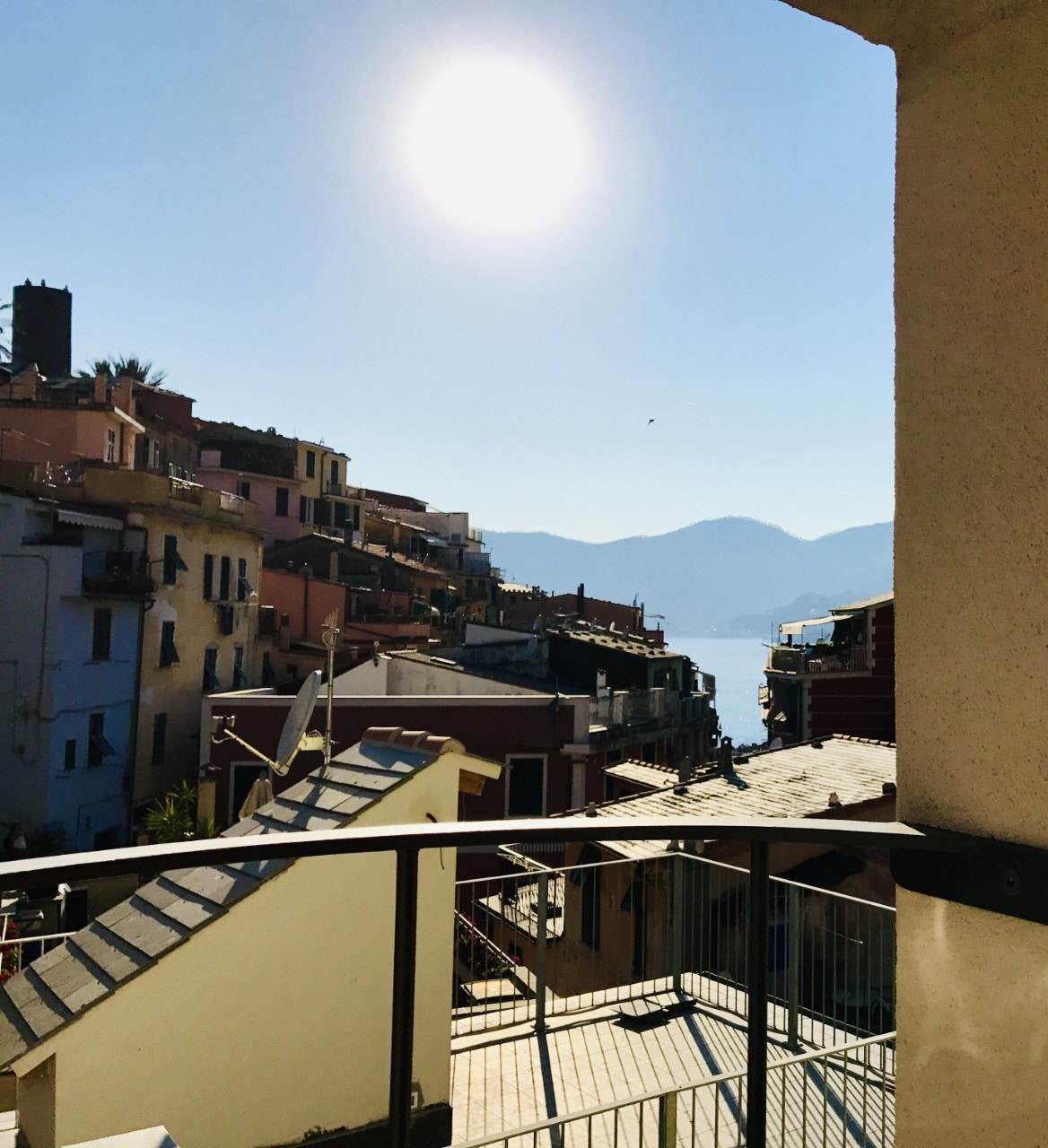 A view from a balcony overlooks a vibrant Mediterranean village with colorful buildings nestled on a hillside. The sun shines brightly in the clear sky, reflecting off the sea in the distance, while the sloped rooftops of the village create an engaging perspective.