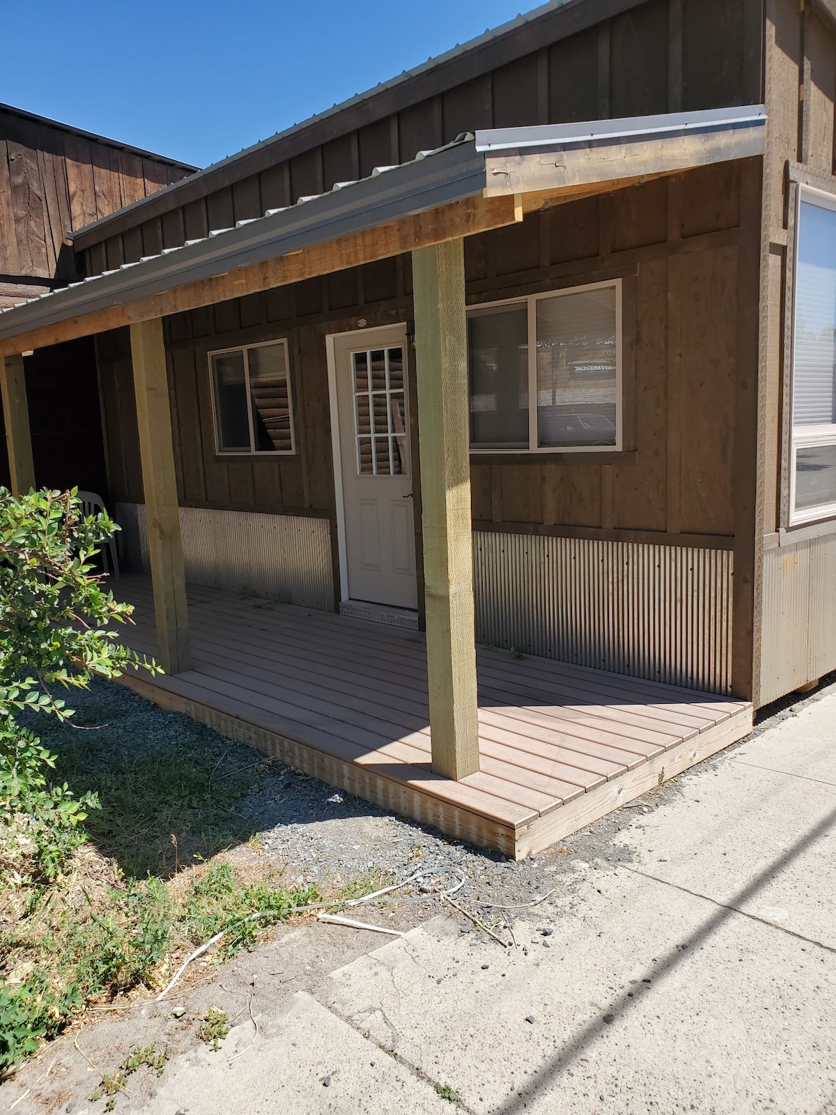 The exterior of the cabin features a covered porch with wooden decking and a panelled door. Large windows allow natural light into the space, while the rustic design complements the surroundings. Greenery borders the pathway leading to the entrance.