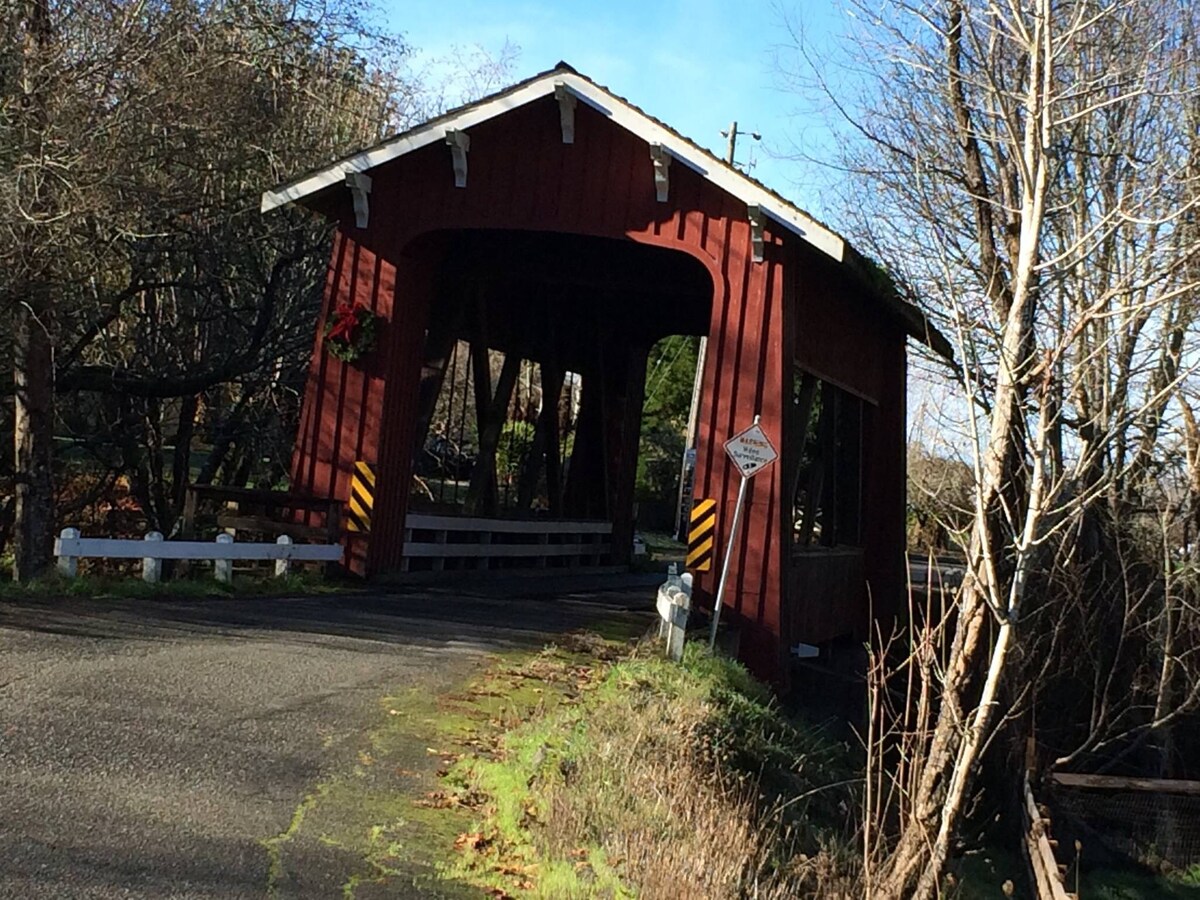 A charming covered bridge is depicted with a red wooden structure, featuring open sides that allow a view of the surrounding greenery. The bridge sits over a quiet road, with seasonal decorations visible, enhancing the serene rural landscape.