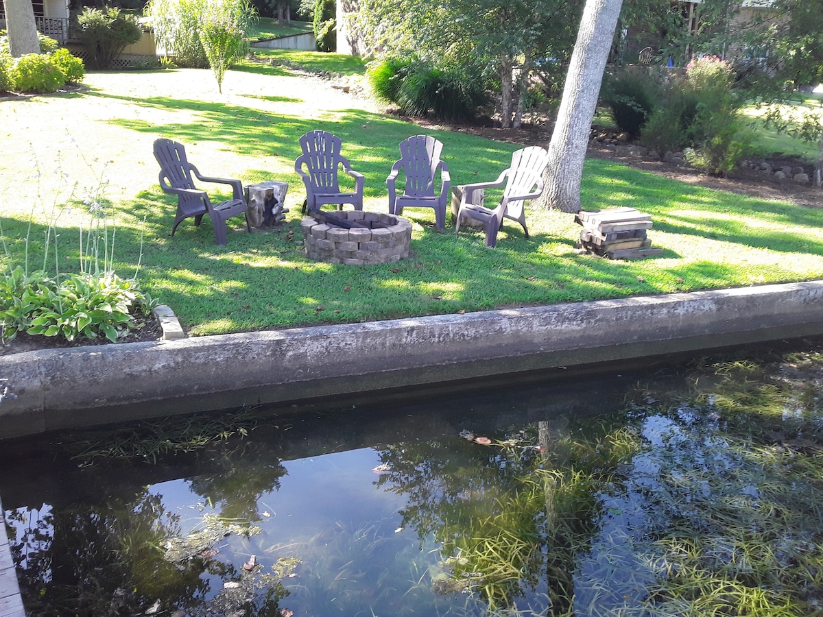 A circular fire pit is surrounded by several Adirondack chairs on a manicured lawn. The area is adjacent to a calm body of water, with reflections of nearby vegetation captured in the water's surface.