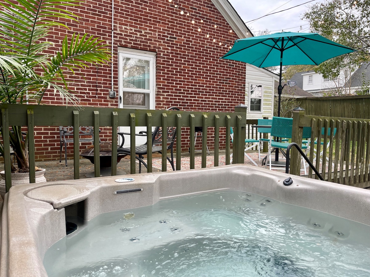 A hot tub is positioned on a wooden deck surrounded by green railings. Nearby, a dining table with chairs and an umbrella adds to the outdoor space. A brick wall in the background provides an inviting visual element.