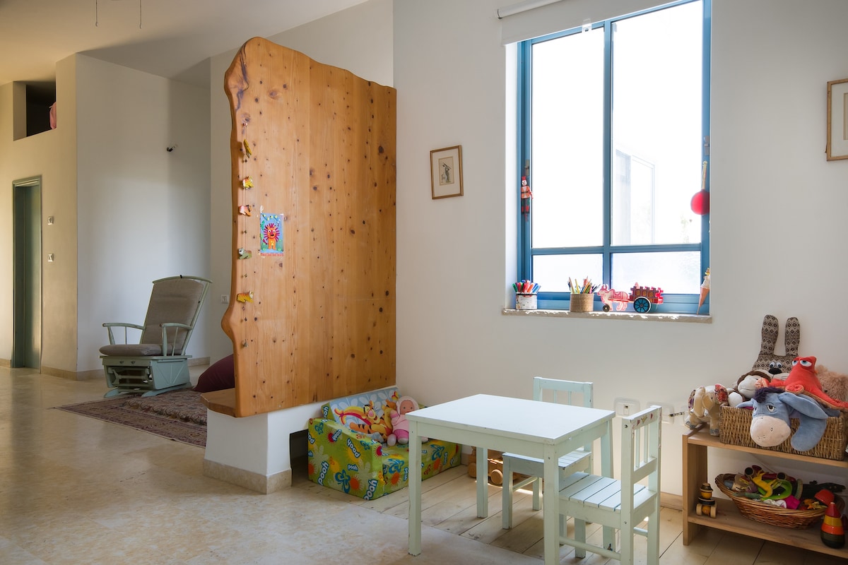 A bright and cheerful play area features a small table with two light-colored chairs. Toys are neatly organized in a colorful storage box, while a wooden divider with decorative accents adds warmth to the space. Natural light flows through a large window, enhancing the inviting atmosphere.