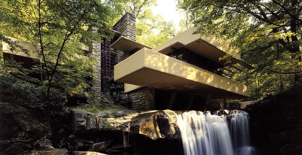 A modern structure is nestled within a lush forest, featuring clean lines and a cantilevered design over a cascading waterfall. Natural stone and concrete elements harmonize with the surrounding greenery, enhancing the serene outdoor setting.