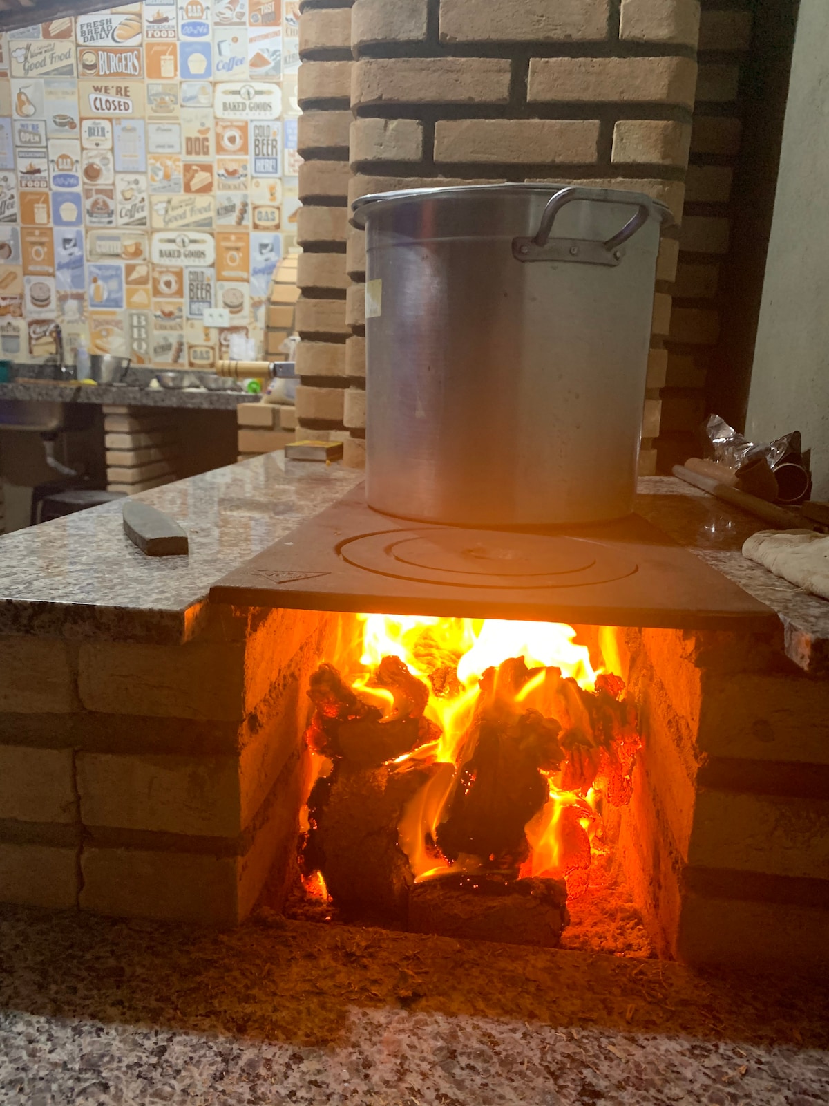 A stove surrounded by stacked bricks features an active flame, with logs burning inside. A large metal pot rests atop the stovetop, reflecting a warm ambiance from the flickering fire. The backdrop reveals a patterned wall, suggesting a cozy cooking area.