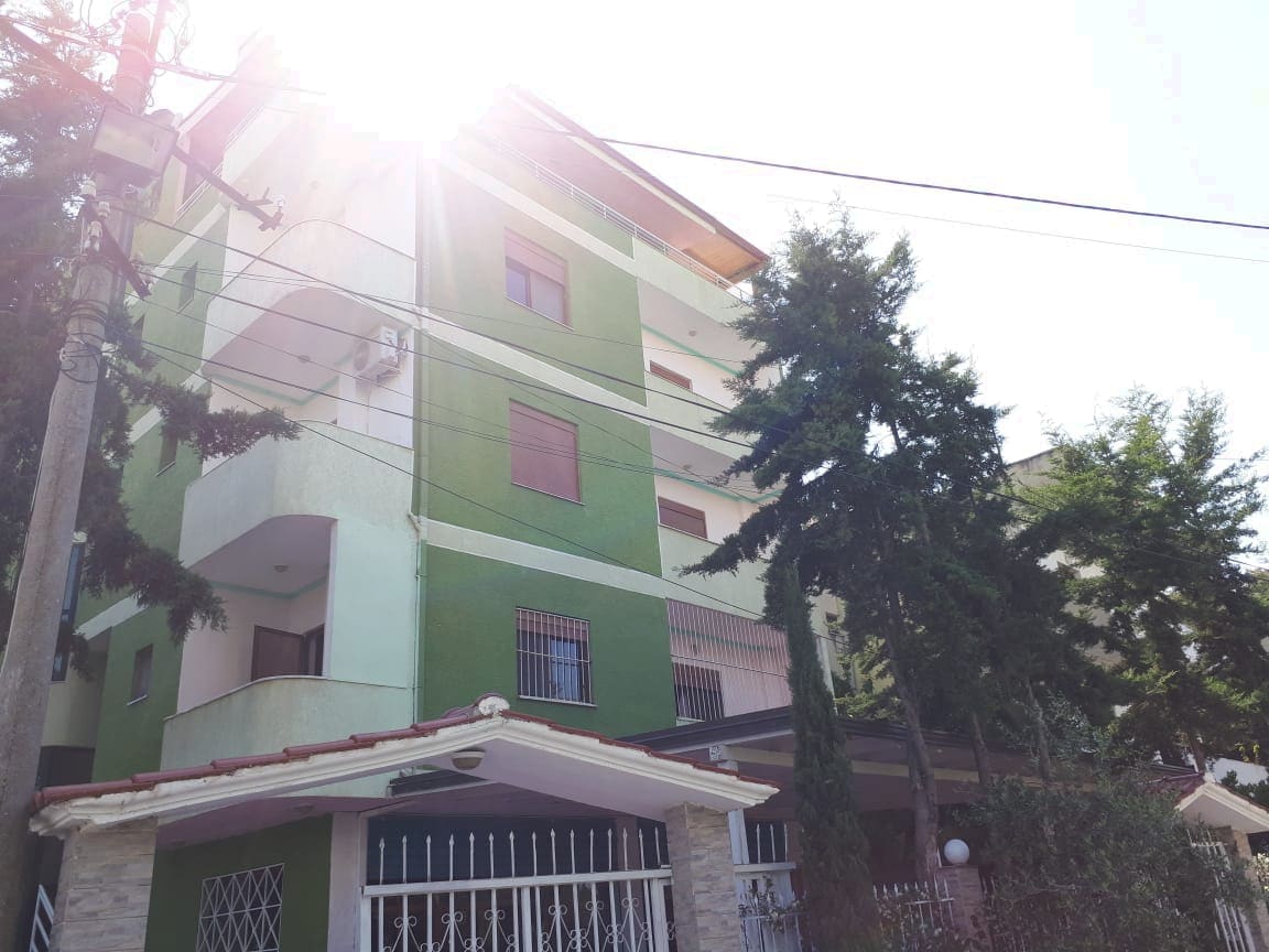 A five-story residential building is depicted, featuring a vibrant green exterior. Sunlight casts a bright glow across the top floors, while lush trees border the property, providing a natural setting. A covered entrance is visible, adding convenience and charm to the setting.