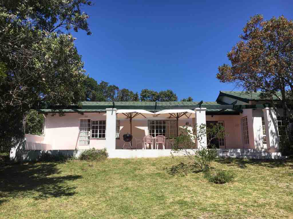 The cottage is set amid greenery, featuring a spacious verandah with outdoor furniture. Light-colored walls contrast with the lush surroundings, while a covered area provides shade. Tall trees frame the property, and the sky is clear, indicating a bright day.