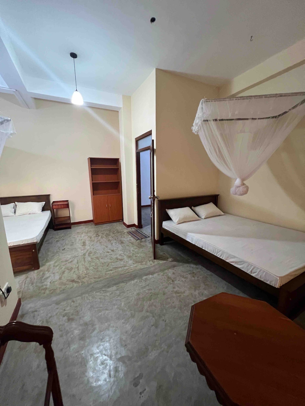 A spacious family room is presented, featuring two queen-size beds and a single bed. The room is furnished with a wardrobe and a small table with chairs. Natural light enters through a beachside window, complemented by a mosquito net over the beds.