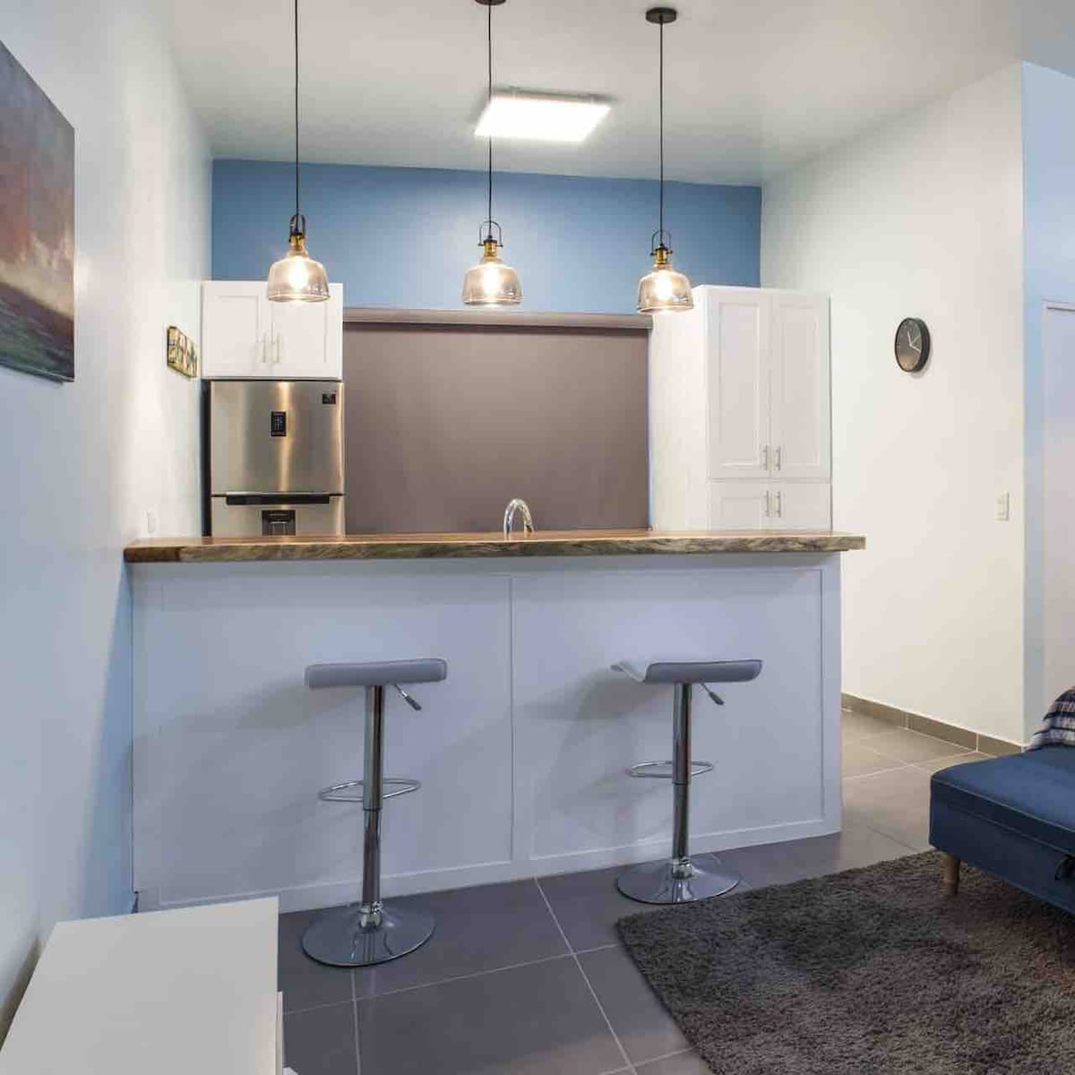 A modern kitchen area is depicted, featuring a sleek bar with two high stools. Above, three pendant lights provide illumination. The kitchen is equipped with stainless steel appliances, including a refrigerator, and the countertops showcase a warm wooden finish.