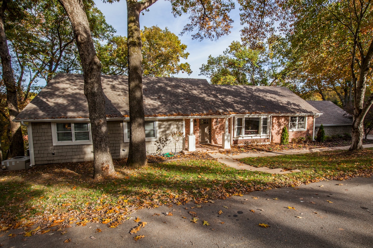 The exterior of a recently remodeled home is shown, featuring a single-story structure with a mix of brick and siding. The surrounding area is adorned with autumn leaves, and large trees provide shade, creating a serene and inviting entrance.