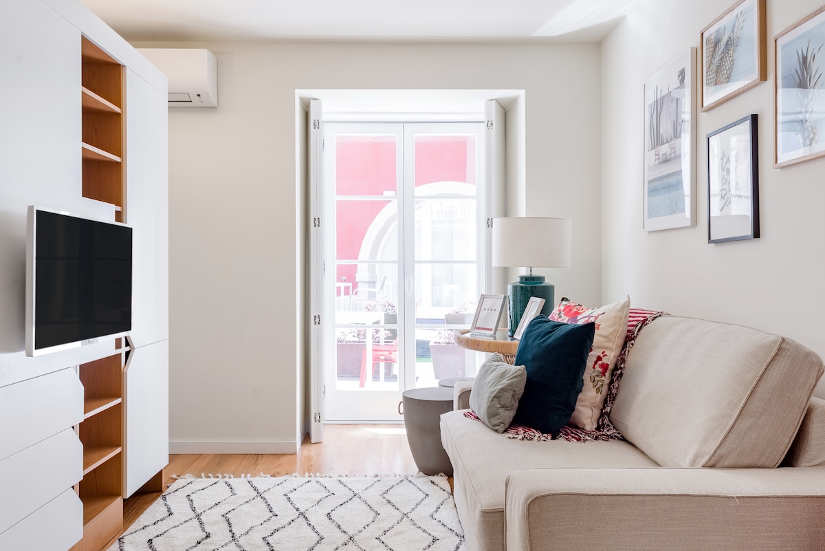 A cozy living area features a light-colored sofa adorned with colorful cushions. A wall-mounted television is positioned above a wooden console. Natural light illuminates the space through large double doors that lead to an outdoor area, enhancing the bright atmosphere.