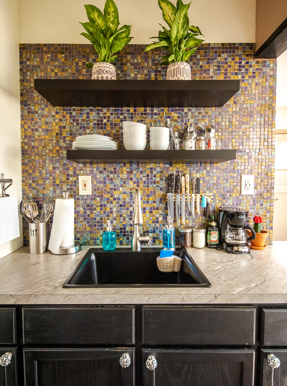 A kitchen area is highlighted by a mosaic tile backsplash in vibrant colors. Above the sink, two shelves display potted plants and dinnerware, while a range of glassware and utensils is organized below. A coffee maker and additional items are visible on the countertop.