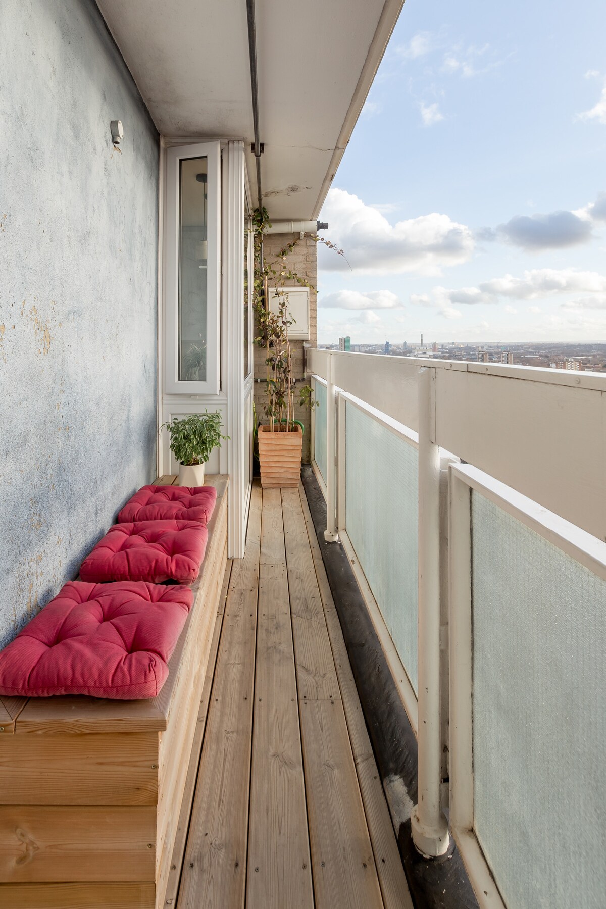 A balcony with wooden flooring features a long built-in seating area adorned with red cushions. Potted plants are positioned at either end, and glass railings offer unobstructed views of the cityscape beyond.