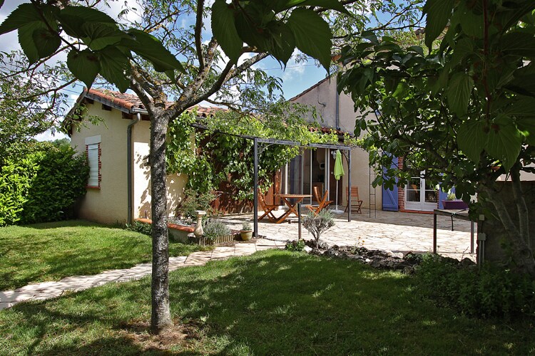 A tranquil outdoor space is surrounded by lush greenery, featuring a stone terrace and comfortable lounge chairs. A pergola provides shade, while a variety of plants add natural beauty. The exterior of the home is visible in the background, with colorful shutters enhancing the ambiance.