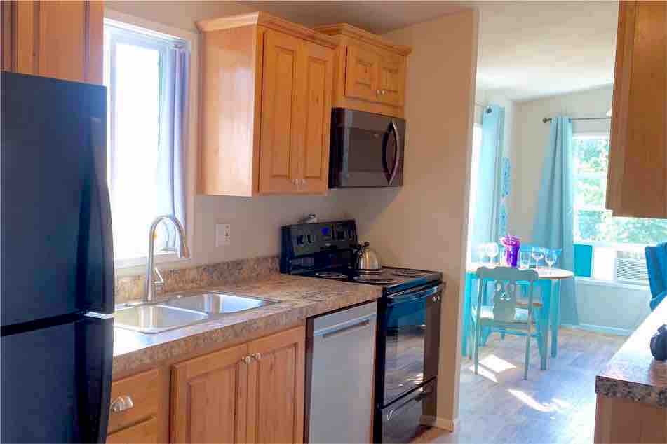 The kitchen features light wood cabinetry and a granite countertop. A stainless-steel stove, microwave, and refrigerator are visible. A dual bowl sink with a pull-down sprayer is positioned under a window. In the distance, a dining area with a small table and chairs can be seen.