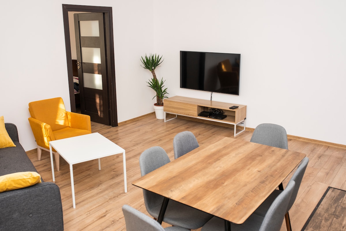 A bright living area features a large flat-screen TV mounted on a light wooden console. A wooden dining table with gray chairs is positioned near a white coffee table, complemented by a yellow armchair. Light wood flooring adds warmth to the space.