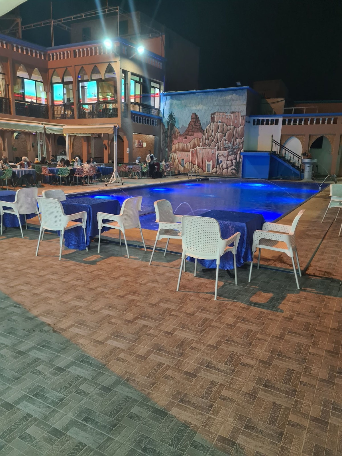 A spacious outdoor area features a large pool illuminated in blue, surrounded by several white chairs arranged neatly around its perimeter. A mural depicting a mountainous landscape is visible on one wall, enhancing the vibrant atmosphere under the night sky.