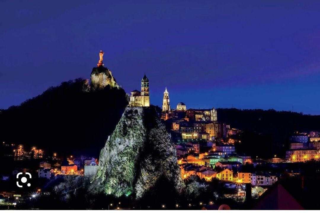 A nighttime view showcases the illuminated landscape of a hillside town, featuring prominent rock formations topped with statues and historic buildings. The warm lights reflect the charm of the area, while the sky transitions from deep blue to darker hues, adding depth to the scene.