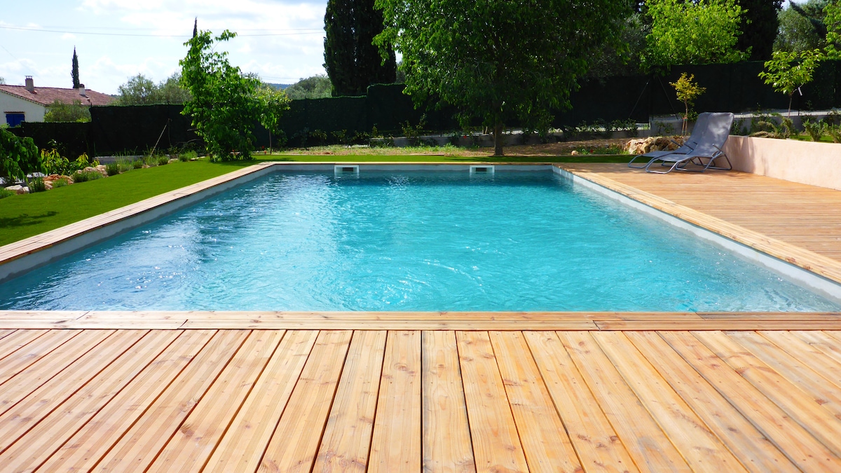 A refreshing swimming pool is nestled within a spacious wooden deck, surrounded by lush greenery. Sunlight glistens off the water's surface, while a lounge chair rests nearby, inviting relaxation. The tranquil setting suggests a serene escape in a private outdoor area.