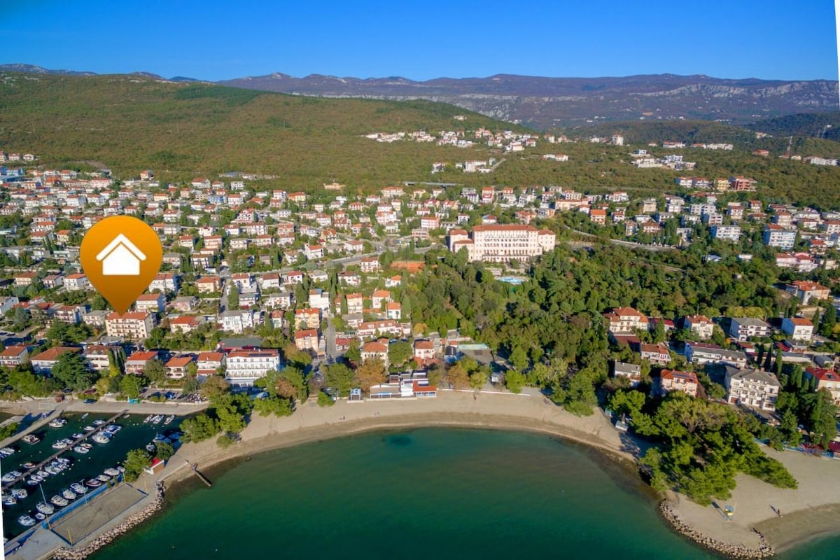 An aerial view captures a coastal area, featuring a sandy beach along a curved shoreline. Residential buildings are visible in the background, along with green hills rising in the distance. The marina hosts a collection of boats, enhancing the serene landscape.