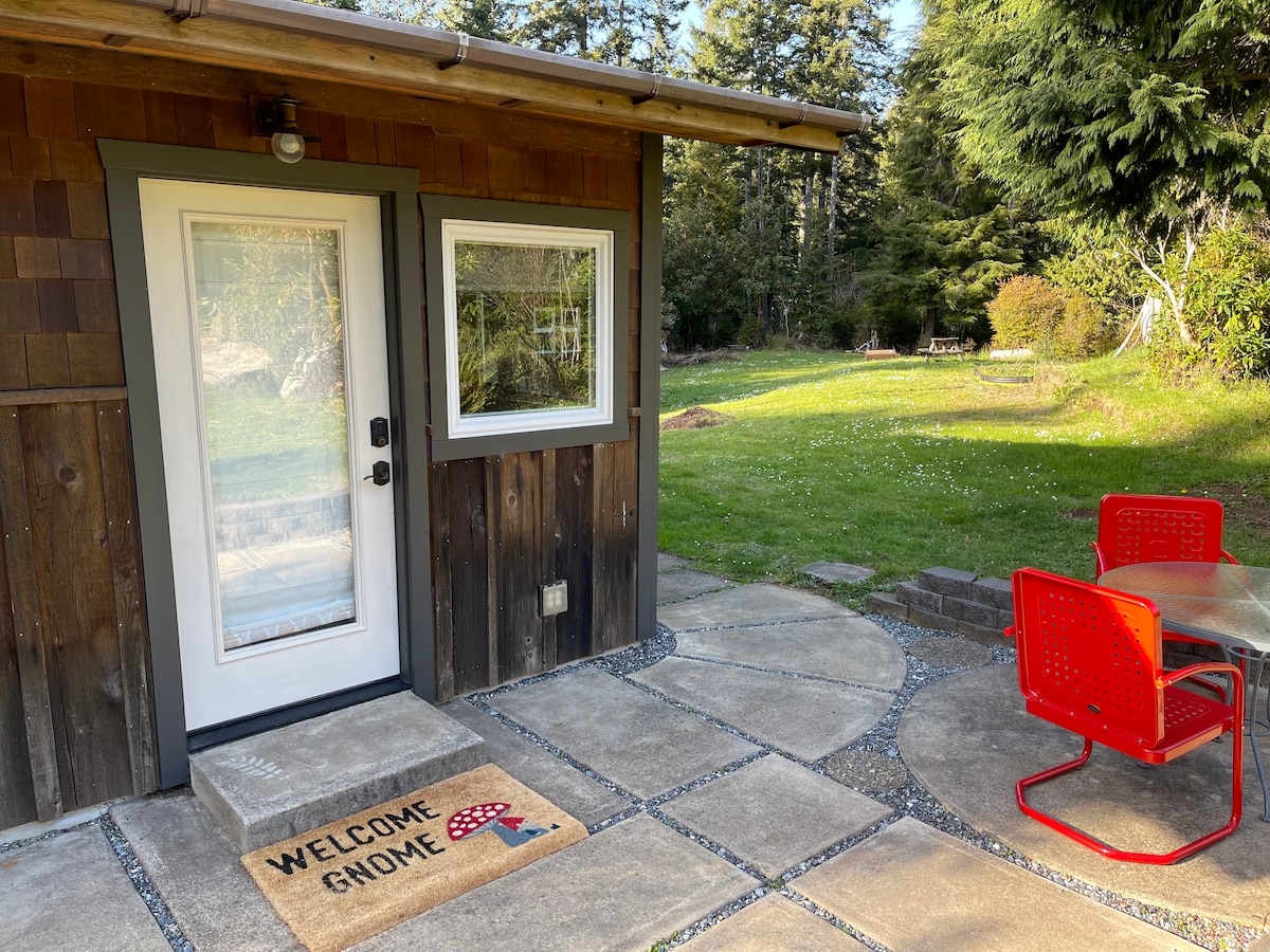 The entrance to the cottage features a decorative welcome mat, with a small patio area that has a round table and two red chairs. Lush greenery and a grassy area surround the entrance, enhancing the natural setting.