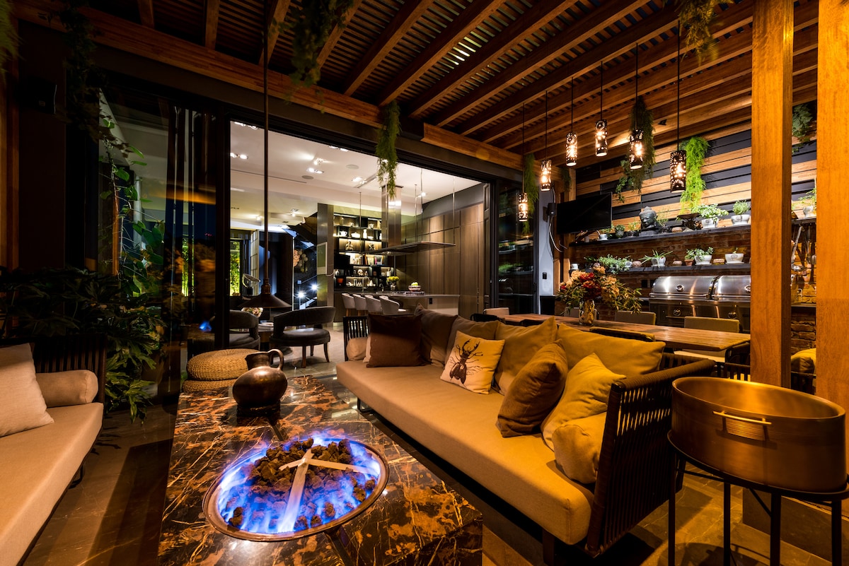 An elegant lounge area features a modern seating arrangement with plush cushions, centered around a striking blue flame fire pit. The space is adorned with hanging plants and warm lighting, creating a cozy ambiance that invites relaxation and socializing. A glimpse of a gourmet kitchen can be seen in the background.