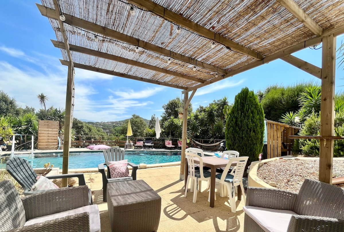 An outdoor area features a shaded seating space under a wooden pergola, with modern wicker furniture arranged around a table. The shared pool is visible in the background, surrounded by lush greenery and lounge chairs, offering a tranquil setting for relaxation.